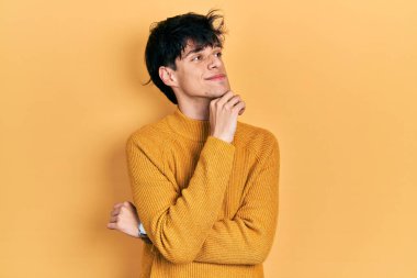 Yakışıklı hippi genç adam sıradan sarı süveter giyiyor, elleri çenesinde soru sormayı düşünüyor, düşünceli bir ifade. Gülümseyen ve düşünceli bir yüz. şüphe kavramı. 