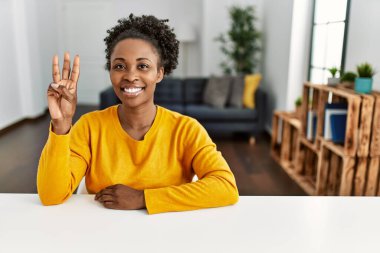 Genç Afro-Amerikalı kadın, gündelik giysiler giyip evdeki masada oturuyor ve üç numarayı göstererek gülümsüyor ve mutlu.. 