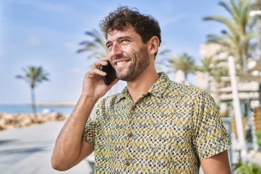 Genç İspanyol adam gülümsüyor. Deniz kenarında akıllı telefondan konuşuyor.