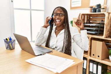 Ofiste çalışan örgülü siyahi kadın telefonda konuşurken şaşkın şaşkın ve şaşkın bir şekilde parmaklarını kaldırıp kollarını kaldırdı.. 
