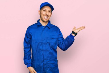 Bald man with beard wearing builder jumpsuit uniform smiling cheerful presenting and pointing with palm of hand looking at the camera. 