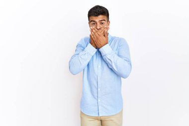 Young hispanic man wearing business shirt standing over isolated background shocked covering mouth with hands for mistake. secret concept. 