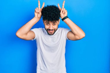 Young arab man with beard wearing casual white t shirt posing funny and crazy with fingers on head as bunny ears, smiling cheerful 