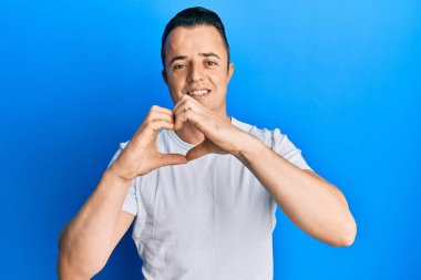 Yakışıklı, beyaz tişört giyen, aşık bir şekilde gülümseyen, elleriyle kalp sembolü yapan genç bir adam. romantik kavram. 