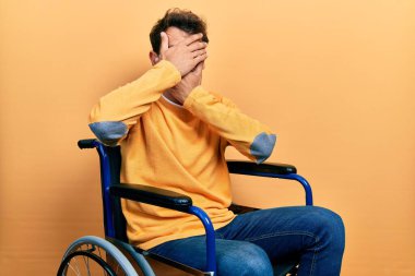 Handsome man with beard sitting on wheelchair covering eyes and mouth with hands, surprised and shocked. hiding emotion 