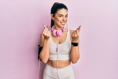 Spor kıyafetleri giyen ve kulaklık takan genç İspanyol bir kadın. Elleriyle para hareketi yapıyor. Maaş istiyor. 