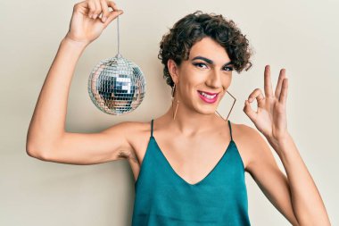 Young hispanic gay holding shiny disco ball doing ok sign with fingers, smiling friendly gesturing excellent symbol 