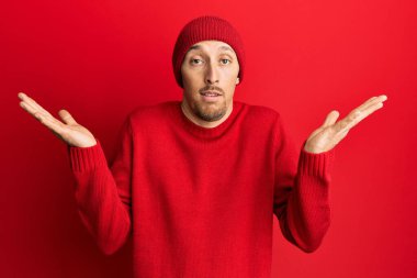 Bald man with beard wearing wool sweater and winter hat clueless and confused with open arms, no idea and doubtful face. 