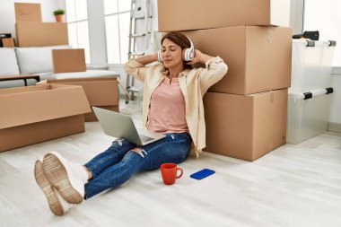 Orta yaşlı beyaz kadın dizüstü bilgisayar ve kulaklık kullanıyor. Yeni evinde yerde oturuyor..