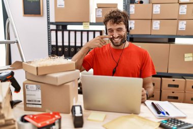 Young hispanic call center agent man working at warehouse pointing with hand finger to face and nose, smiling cheerful. beauty concept 