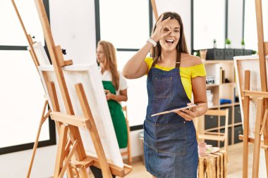 Young hispanic woman at art classroom smiling happy doing ok sign with hand on eye looking through fingers 