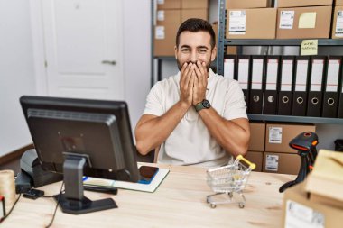 Handsome hispanic man working at small business commerce laughing and embarrassed giggle covering mouth with hands, gossip and scandal concept 