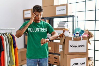 Young handsome hispanic man wearing volunteer t shirt at donations stand looking at the watch time worried, afraid of getting late 