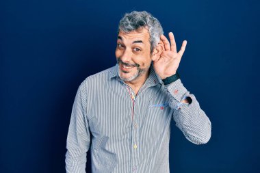 Handsome middle age man with grey hair wearing business shirt smiling with hand over ear listening an hearing to rumor or gossip. deafness concept. 