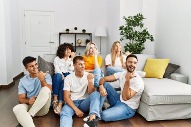 Group of people sitting on the sofa and floor at home smiling with happy face looking and pointing to the side with thumb up. 