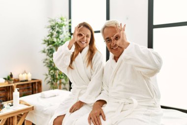 Middle age hispanic couple wearing bathrobe at wellness spa smiling happy doing ok sign with hand on eye looking through fingers 