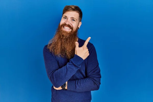 红头发的男人留着长胡子 蓝色背景上穿着休闲的蓝色毛衣 脸上挂着大大的笑容 手指头指向镜头的侧面 — 图库照片