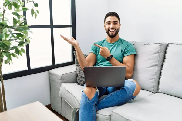 手で提示し 指で指している間 自宅でソファの上に座って驚きとカメラに笑みを浮かべてコンピュータのラップトップを使用してひげを持つ若いハンサムな男 — ストック写真