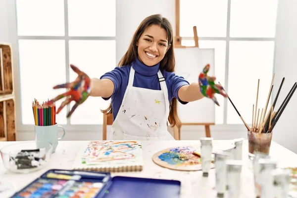 Young brunette woman at art studio with painted hands looking at the camera smiling with open arms for hug. cheerful expression embracing happiness.