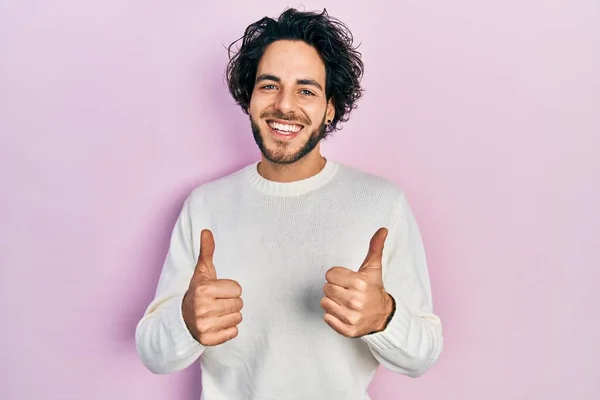 手で積極的なジェスチャーを行うカジュアルな白いセーターの成功サインを身に着けているハンサムなヒスパニック系の男 親指を笑顔と幸せ 陽気な表情と勝者のジェスチャー — ストック写真