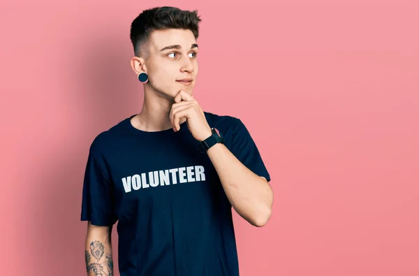 耳の拡張機能を持つ若い白人の少年は 集中的な表現について考えて顎に手でボランティアのTシャツを着て 笑顔で思いやりのある顔 疑わしい概念 — ストック写真