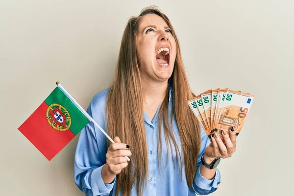 ポルトガル国旗とユーロ紙幣を持っている若いブロンドの女性怒っていると怒っている叫び不満と激怒 怒りで叫んで検索アップ — ストック写真