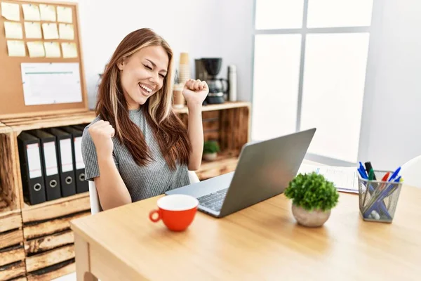 Young brunette woman working at the office with laptop very happy and excited doing winner gesture with arms raised, smiling and screaming for success. celebration concept. 