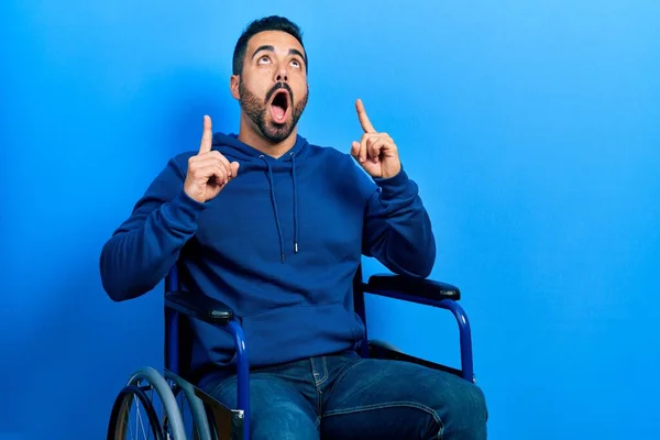 Handsome Hispanic Man Beard Sitting Wheelchair Amazed Surprised Looking Pointing — Fotografia de Stock