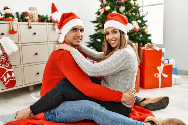 Młoda Para Uśmiechnięta Szczęśliwa Świątecznym Kapeluszu Siedząc Podłodze Przytulając Się — Zdjęcie stockowe