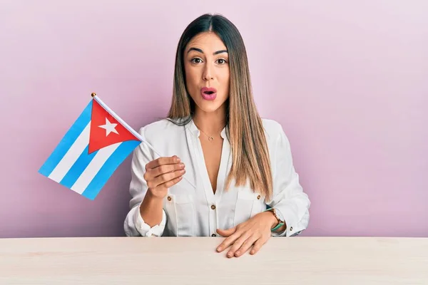 若いヒスパニック系の女性キューバ国旗を持ってテーブルの上に座って怖がって驚きのために口を開けて驚いて 不信の顔 — ストック写真