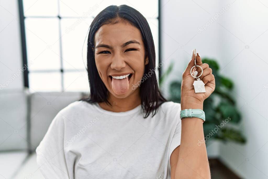 Young hispanic woman holding keys of new home sticking tongue out happy with funny expression. emotion concept. 