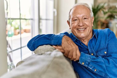 Evinin oturma odasındaki kanepede oturan gri saçlı son sınıf öğrencisi. Evde mutlu gülümseyen olgun bir adam..