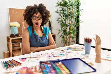Beautiful african american woman with afro hair painting at art studio afraid and shocked with surprise expression, fear and excited face. 