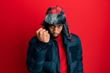Young african american man with beard wearing winter hat and coat angry and mad raising fist frustrated and furious while shouting with anger. rage and aggressive concept. 