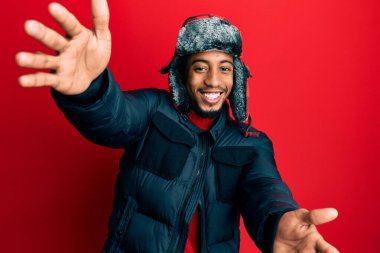 Young african american man with beard wearing winter hat and coat looking at the camera smiling with open arms for hug. cheerful expression embracing happiness. 