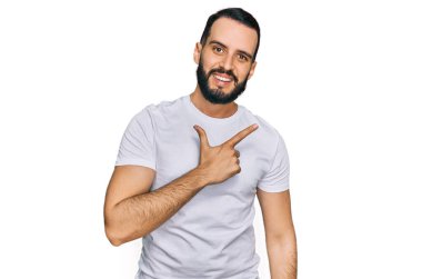 Young man with beard wearing casual white t shirt cheerful with a smile on face pointing with hand and finger up to the side with happy and natural expression 
