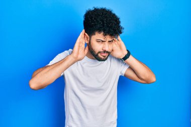 Young arab man with beard wearing casual white t shirt trying to hear both hands on ear gesture, curious for gossip. hearing problem, deaf 