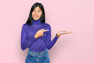 Beautiful young asian woman wearing casual clothes amazed and smiling to the camera while presenting with hand and pointing with finger. 