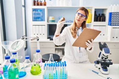 Young blonde woman wearing scientist uniform holding kiwi and clipboard at laboratory