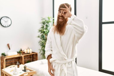 Redhead man with long beard wearing bathrobe at wellness spa smiling happy doing ok sign with hand on eye looking through fingers 