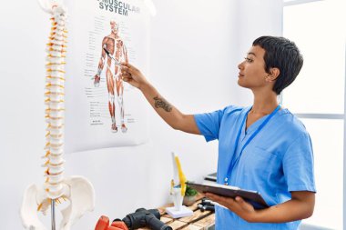 Young hispanic woman wearing physiotherapist uniform pointing to vertebral column holding checklist at physiotherapy clinic