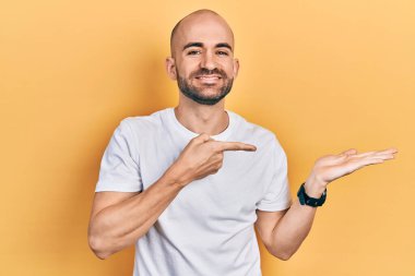Young bald man wearing casual white t shirt amazed and smiling to the camera while presenting with hand and pointing with finger. 