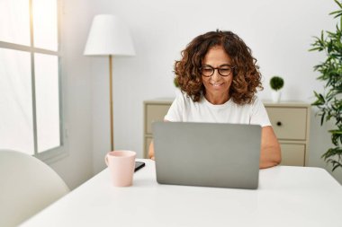 Orta yaşlı İspanyol kadın evde dizüstü bilgisayar kullanarak gülümsüyor.