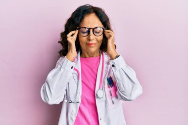 Middle age hispanic woman wearing doctor uniform and glasses with hand on head for pain in head because stress. suffering migraine. 