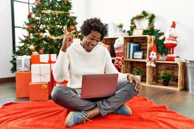 Genç Afro-Amerikan adam dizüstü bilgisayar kullanıyor Noel ağacının yanında oturuyor ve başarılı bir fikir işaret ediyor. Heyecanlı ve mutlu. Bir numara.. 