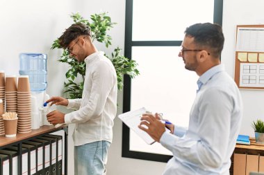Two hispanic men business workers drinking water and writing on clipboard working at office