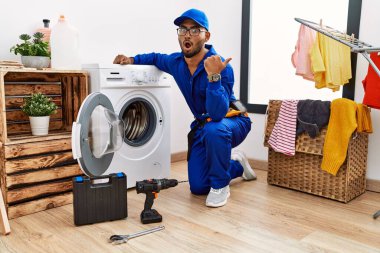 Young indian technician working on washing machine surprised pointing with hand finger to the side, open mouth amazed expression. 