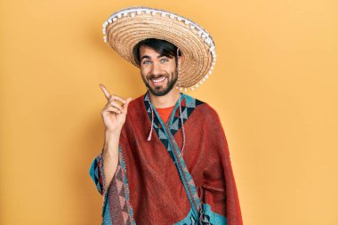Young hispanic man holding mexican hat with a big smile on face, pointing with hand and finger to the side looking at the camera. 