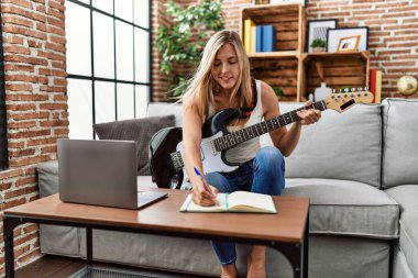 Genç sarışın kadın evde gitar çalarken kendinden emin gülümsüyor.