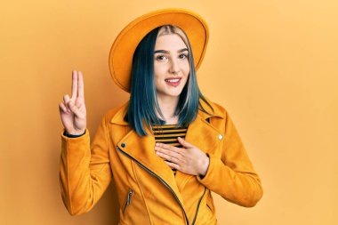 Young modern girl wearing yellow hat and leather jacket smiling swearing with hand on chest and fingers up, making a loyalty promise oath 